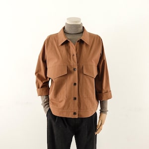 Può includere: Una giacca marrone abbottonata con colletto, due ampie tasche sul petto e maniche a tre quarti, indossata sopra un maglione a collo alto grigio a coste e abbinata a pantaloni grigio scuro su un manichino.