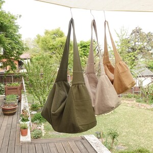 May include: Three hobo-style shoulder bags hanging in a row, featuring a deep olive green bag in the foreground, followed by a light beige bag and a tan bag. Each bag has a wide shoulder strap, a slouchy body, and a visible side pocket.