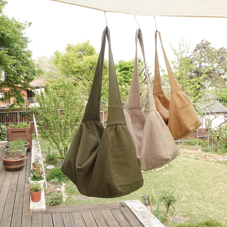 May include: Three hobo-style shoulder bags hanging in a row, featuring a deep olive green bag in the foreground, followed by a light beige bag and a tan bag. Each bag has a wide shoulder strap, a slouchy body, and a visible side pocket.