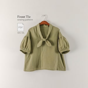 Puede incluir: Una blusa verde salvia con mangas abullonadas hasta el codo, escote en V y un lazo frontal. El texto "Front Tie sewing pattern" y "PDF XXS-5XL" es visible. La prenda se muestra colgada en una percha sobre un fondo neutro.