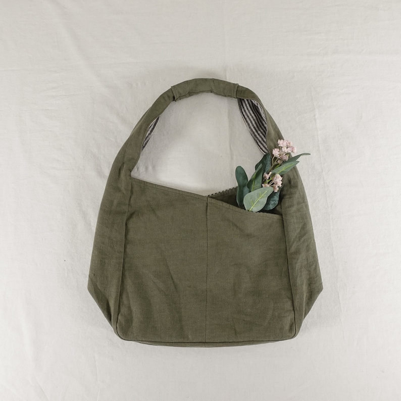 May include: An olive green hobo bag with a single shoulder strap and a front pocket. The interior of the strap features a black and white striped pattern. A small sprig of artificial green leaves with tiny pink flowers is tucked into the pocket.