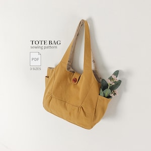 Op de afbeelding: Een mosterdgele tote bag met twee schouderbanden, een centrale knoopsluiting en zijzakken. De tekst TOTE BAG sewing pattern PDF 3 SIZES is zichtbaar. De binnenvoering heeft een kleine bloemenprint en er zit een groene plant in de tas.