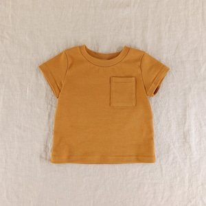 May include: A short-sleeved t-shirt in a solid mustard yellow colour. The garment features a crew neckline and a single patch pocket located on the left side of the chest. The shirt is constructed with a simple, clean design and a straight hemline.