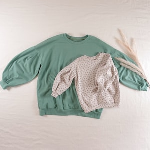 May include: Two sweatshirts, one green and one with a floral pattern, are laid out on a white surface. Both sweatshirts have puffed sleeves.
