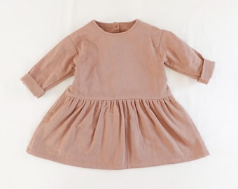 May include: A long-sleeved dress in a solid dusty rose color. The garment features a round neckline, a drop-waist seam, and a gathered skirt. The sleeves are finished with folded cuffs, and the back of the neckline includes a single button closure.