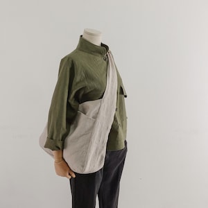 May include: An olive green long-sleeved button-up shirt with a high collar, paired with dark gray trousers and a large, light beige slouchy crossbody bag made of a textured fabric, worn over the shoulder across the torso.