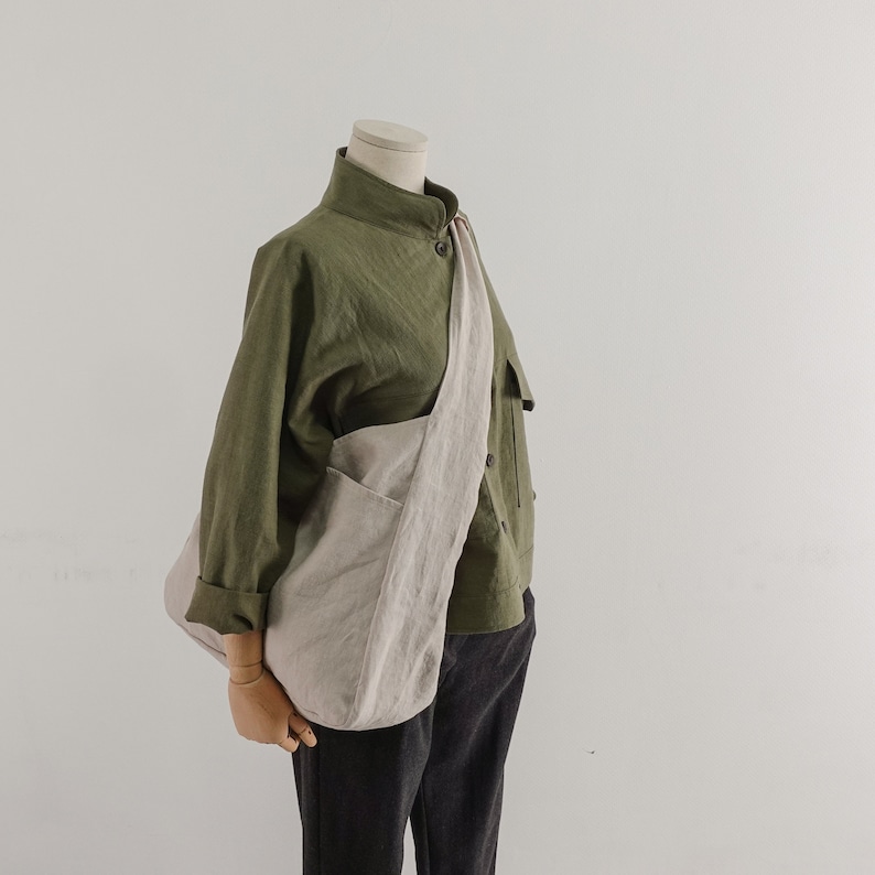 May include: An olive green long-sleeved button-up shirt with a high collar, paired with dark gray trousers and a large, light beige slouchy crossbody bag made of a textured fabric, worn over the shoulder across the torso.
