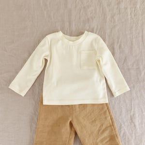 May include: A long-sleeved cream-coloured shirt with a single chest pocket and a crew neckline, paired with tan-coloured trousers. The shirt features a simple design with long sleeves and a straight hem, while the trousers have a visible fabric texture.