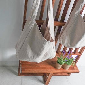 May include: Two beige hobo-style shoulder bags made of a textured fabric, featuring a wide strap and a deep, open main compartment. The bags are hanging from a wooden rack, with two small potted lavender plants placed on the shelf below them.