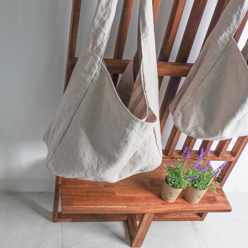 May include: Two beige hobo-style shoulder bags made of a textured fabric, featuring a wide strap and a deep, open main compartment. The bags are hanging from a wooden rack, with two small potted lavender plants placed on the shelf below them.
