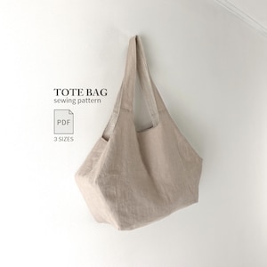 Op de afbeelding: Een beige tote bag met twee schouderbanden, hangend tegen een effen muur. De tekst TOTE BAG sewing pattern, een PDF-pictogram en de woorden 3 SIZES staan links van de tas, wat aangeeft dat het een digitaal naaipatroon betreft.