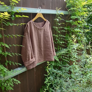 Op de afbeelding: Een bruin linnen top met een ronde hals en lange mouwen. De top hangt aan een houten hanger tegen een houten schutting.