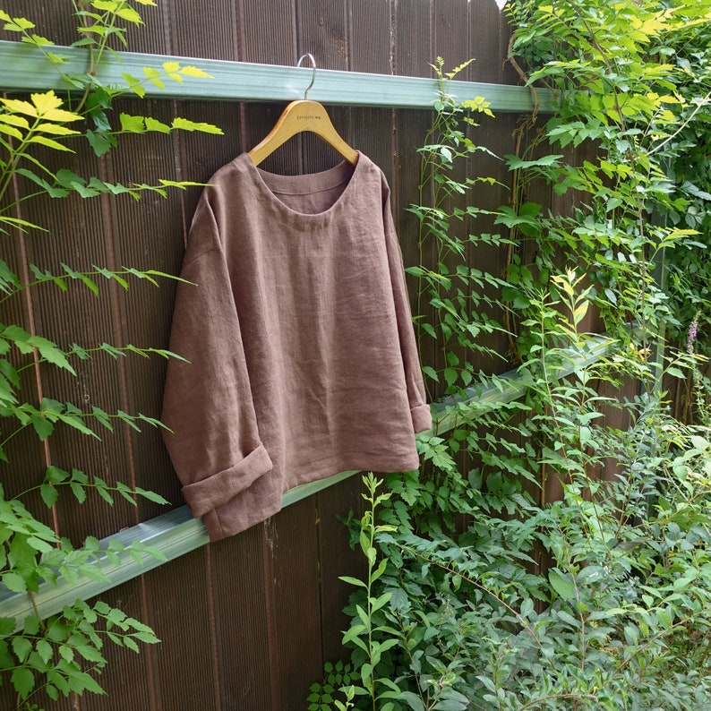 Op de afbeelding: Een bruin linnen top met een ronde hals en lange mouwen. De top hangt aan een houten hanger tegen een houten schutting.