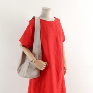 May include: A bright red short-sleeved dress with a round neckline worn with a light beige hobo-style shoulder bag. The bag features a wide strap and a soft, unstructured shape, resting against the side of the dress on a mannequin torso.