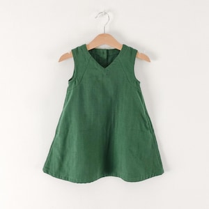 Puede incluir: Un vestido sin mangas de color verde bosque con escote en V y cierre de botones en la espalda. El vestido presenta una silueta sencilla en forma de A con bolsillos laterales visibles y se muestra en una percha de madera.