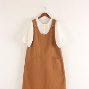 Könnte beinhalten: Ein braunes ärmelloses Trägerkleid mit Rundhalsausschnitt und zwei Vordertaschen, kombiniert mit einer weißen kurzärmeligen Bluse, die eine durchgehende Knopfleiste und vertikale Falten aufweist. Das Kleidungsstück hängt an einem Holzkleiderbügel.