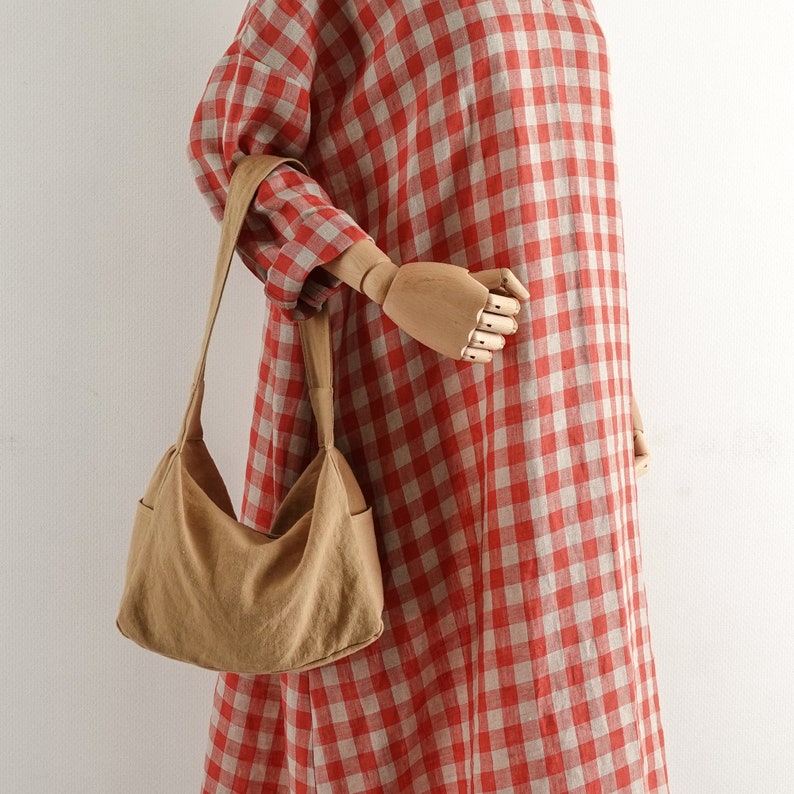 May include: A tan linen tote bag with a long strap. The bag is being held by a wooden hand model wearing a red and white gingham dress.