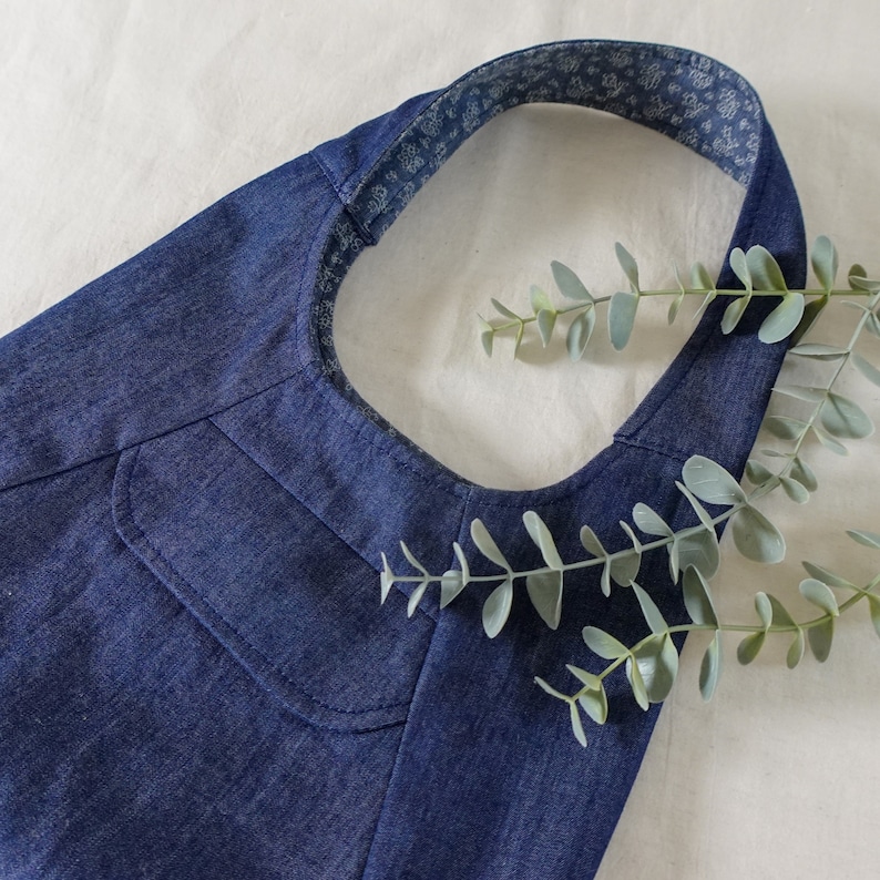 May include: A blue denim tote bag with a white floral patterned strap. The bag has a single pocket on the front.