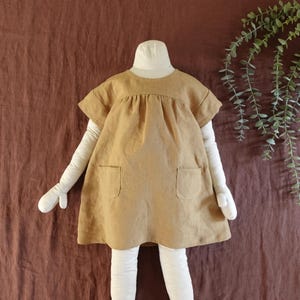 May include: A short-sleeved, tan-coloured dress featuring a round neckline, a gathered bodice, and two square patch pockets on the front. The garment is displayed on a mannequin with white arms and legs against a dark brown fabric background.