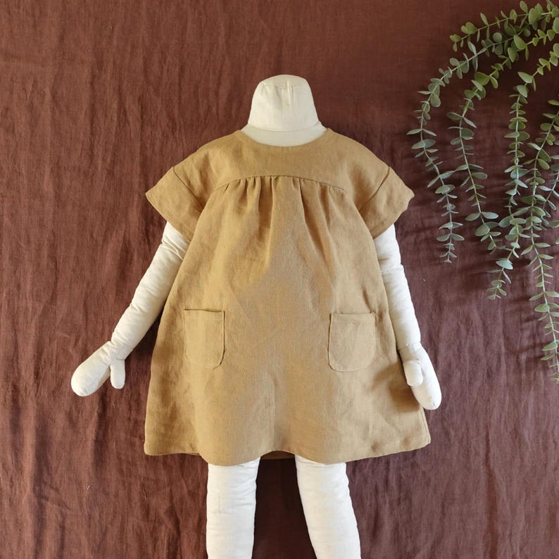 May include: A short-sleeved, tan-coloured dress featuring a round neckline, a gathered bodice, and two square patch pockets on the front. The garment is displayed on a mannequin with white arms and legs against a dark brown fabric background.