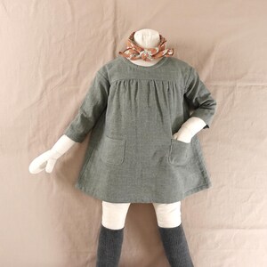 May include: A long-sleeved olive green dress with a round neckline, gathered bodice, and two front patch pockets. The dress is paired with a rust-coloured patterned neck scarf and dark grey ribbed leggings on a mannequin form.