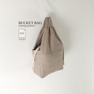 May include: A beige bucket bag sewing pattern featuring a drawstring closure with thick cream-colored rope cords. The text BUCKET BAG sewing pattern, PDF, and 3 SIZES is printed in dark gray on the left side of the bag.