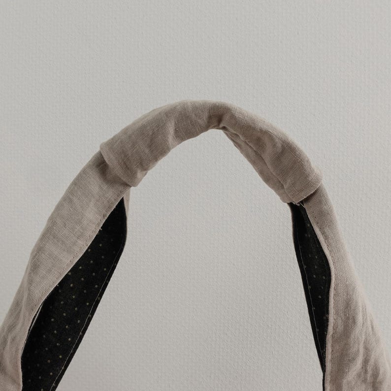 May include: A beige fabric headband with a knotted top detail. The exterior is a solid light brown linen-like material, while the interior lining is a dark charcoal fabric featuring a pattern of small, evenly spaced white polka dots.