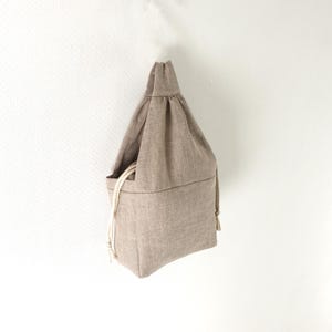 Puede incluir: Una bolsa con cordón de color marrón claro hecha de tela texturizada. La bolsa presenta una sección superior fruncida con un asa única y dos cordones de color blanco roto en los laterales que cierran la abertura, creando una forma estructurada.
