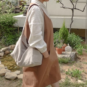 May include: A woman wearing a brown linen dress with a white linen shirt underneath. She is carrying a large, light gray tote bag over her shoulder. The bag has a rectangular shape with a curved top.