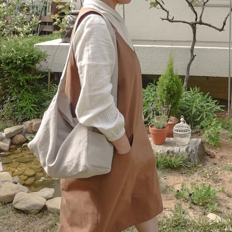 May include: A woman wearing a brown linen dress with a white linen shirt underneath. She is carrying a large, light gray tote bag over her shoulder. The bag has a rectangular shape with a curved top.