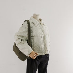 May include: A cream-colored quilted jacket with a diamond pattern, a collared neckline, and a front button closure. The jacket features two large patch pockets on the front and is paired with a dark olive green shoulder bag and dark trousers.