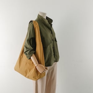 May include: A brown tote bag with a long strap, worn over a green button-up shirt and beige pants.