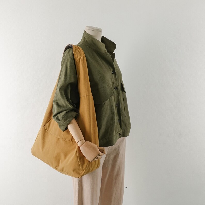 May include: A brown tote bag with a long strap, worn over a green button-up shirt and beige pants.