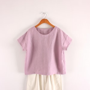 May include: A short-sleeved, round-neck top in a soft lavender color, featuring a relaxed fit and a straight hemline. The garment is displayed on a wooden hanger, positioned above a pair of off-white trousers with a visible vertical seam.