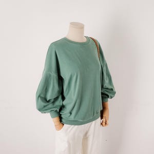 Könnte beinhalten: Ein salbeigrünes Langarm-Sweatshirt mit Rundhalsausschnitt und voluminösen Puffärmeln mit eng anliegenden Bündchen. Das Kleidungsstück wird auf einem Schaufensterpuppen-Torso präsentiert und zeigt ein einfarbiges Design mit lockerer Passform und geripptem Saum.