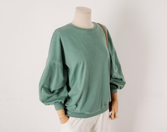 May include: A sage green long-sleeve sweatshirt featuring a crew neckline and voluminous puff sleeves with fitted cuffs. The garment is displayed on a mannequin torso, showcasing a solid color design with a relaxed fit and a ribbed hemline.