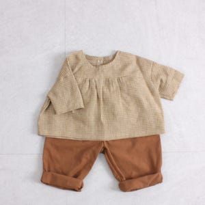 May include: A beige and white gingham short-sleeved top with a round neckline and gathered detailing at the chest, paired with solid brown shorts featuring cuffed hems. The outfit consists of a loose-fitting blouse and matching pull-on style shorts.