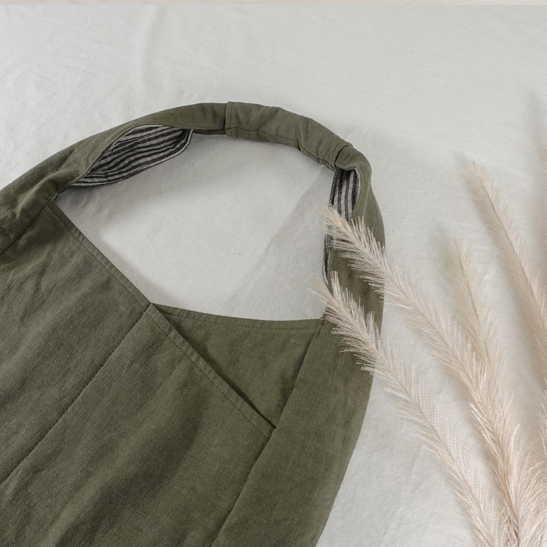 May include: An olive green hobo bag featuring a wide, integrated shoulder strap. The interior of the strap is lined with a black and white striped fabric. The exterior of the bag has a visible seam running vertically down the center of the front panel.