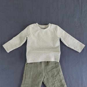 May include: A long-sleeve crew neck shirt in a light heather gray color paired with olive green pants. The shirt features a simple round neckline and long sleeves, while the pants have a visible textured fabric and a straight-leg design.