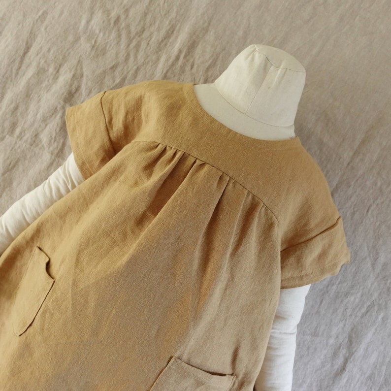 May include: A mustard yellow short-sleeved dress featuring a round neckline, gathered fabric at the chest, and two front pockets. The dress is displayed on a mannequin wearing a long-sleeved white base layer underneath the garment.
