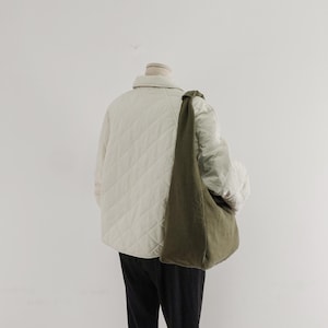 May include: A cream-colored quilted jacket with a collar and long sleeves, worn over black trousers. An olive green hobo-style shoulder bag with a knotted strap detail hangs across the torso. The jacket features a diamond-shaped stitched pattern.