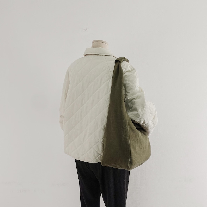 May include: A cream-colored quilted jacket with a collar and long sleeves, worn over black trousers. An olive green hobo-style shoulder bag with a knotted strap detail hangs across the torso. The jacket features a diamond-shaped stitched pattern.