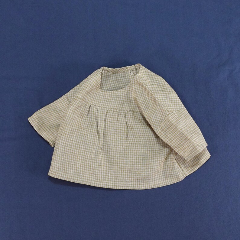 May include: A beige and white gingham check blouse with a round neckline, short sleeves, and a gathered bodice. The garment features a single button closure at the back of the neck and a loose, flowing silhouette with a textured, lightweight fabric.