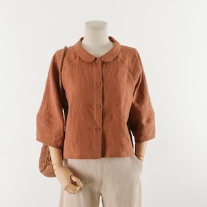 May include: A rust-colored linen shirt with a peter pan collar and button-up front. The shirt has 3/4 sleeves and a relaxed fit.