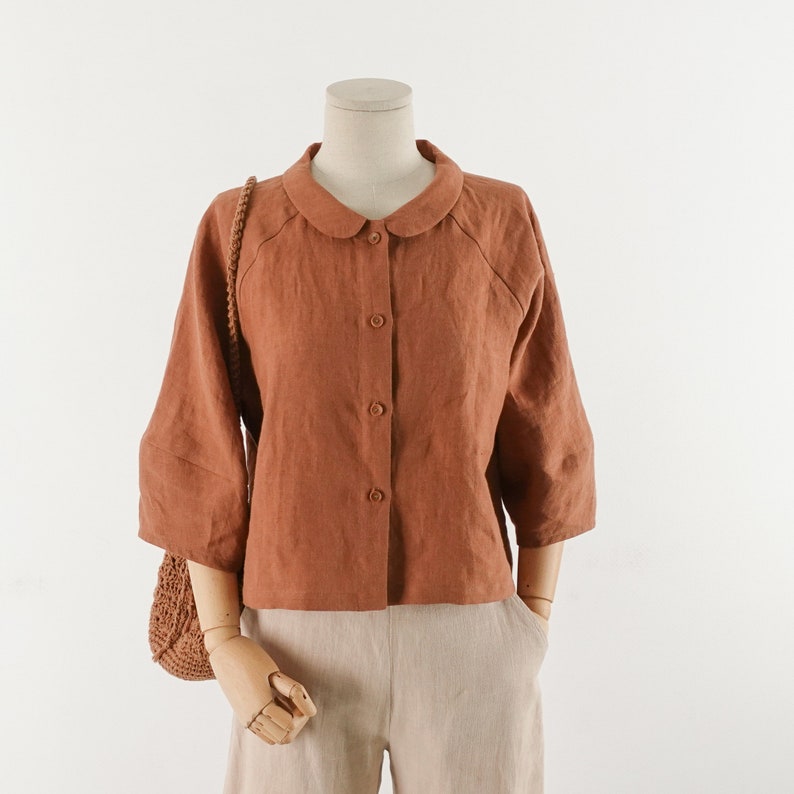 May include: A rust-colored linen shirt with a peter pan collar and button-up front. The shirt has 3/4 sleeves and a relaxed fit.