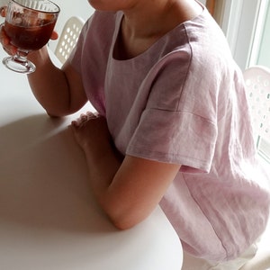 Op de afbeelding: Een vrouw draagt een lichtroze linnen top met een ronde hals en korte mouwen. De top heeft een losse pasvorm en is gemaakt van een lichtgewicht stof.