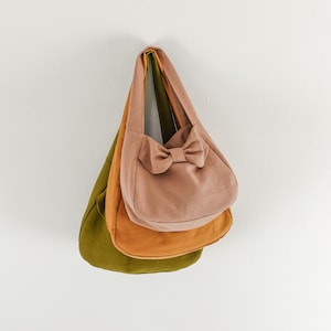 May include: Three stacked hobo bags in olive green, mustard yellow, and dusty rose pink. Each bag features a single shoulder strap and a soft, rounded silhouette. The top dusty rose bag is accented with a prominent fabric bow on the front side.