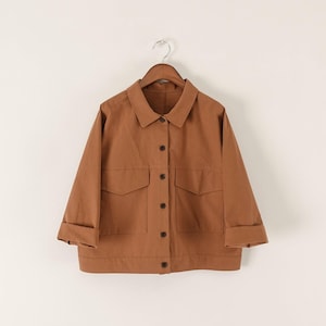 May include: A solid brown button-down jacket featuring a classic collar, long sleeves with folded cuffs, and two large front pockets with flaps. The garment is constructed with a central vertical row of dark brown buttons and a straight hemline.