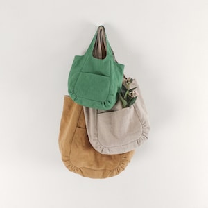 Pode incluir: Três sacos tote em verde, bege e castanho claro, cada um com fundo arredondado, um bolso frontal grande com detalhes franzidos nas bordas e alças integradas. Os sacos estão sobrepostos, com um pequeno ramo verde no saco bege.
