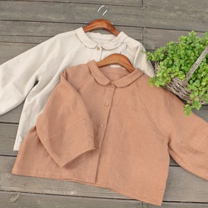 May include: Two linen shirts with peter pan collars, one beige and one brown, hanging on wooden hangers.